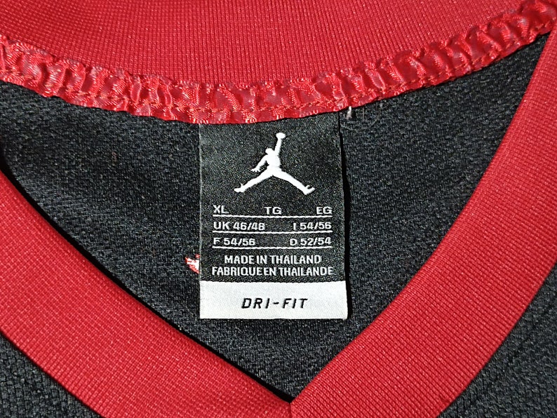 Puede incluir: Camiseta deportiva negra y roja con una etiqueta blanca que dice "XL, UK 46/48, F 54/56, TG, 1 54/56, D 52/54, EG, MADE IN THAILAND, FABRIQUEEN THAILANDE, DRI-FIT".