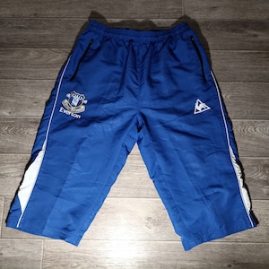 Könnte beinhalten: Blaue Sport-Shorts mit weißen Seitenpanelen und einem weißen Logo am rechten Bein. Das Logo ist ein Hahn in einem Dreieck. Das linke Bein hat ein blaues und goldenes gesticktes Logo mit dem Text "Everton" und einem Wappen.