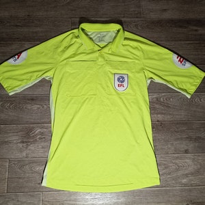 Könnte beinhalten: Ein leuchtend gelbes Schiedsrichtertrikot mit weißen Abschlüssen und einem Aufnäher auf der linken Brust, auf dem "EFL" steht. Das Trikot hat auf jedem Ärmel einen roten und weißen Aufnäher.
