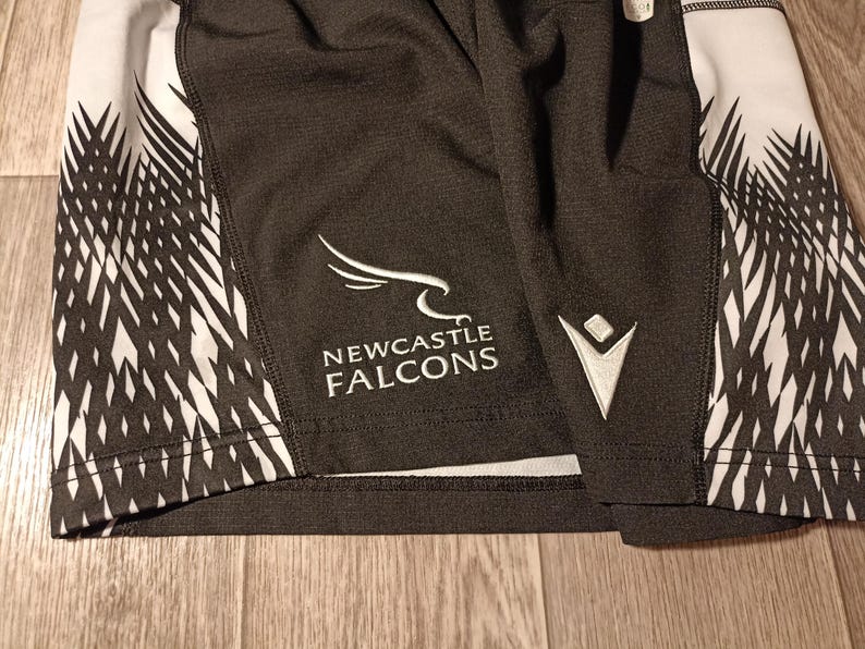 May include: Black and white athletic shorts with a white logo that says "NEWCASTLE FALCONS" and a white chevron design on the side.