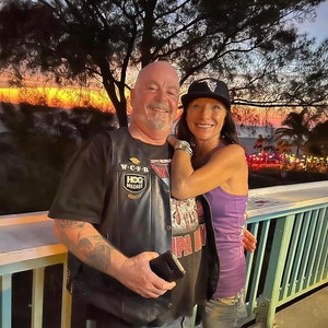 May include: A couple poses for a photo on a balcony overlooking a beach at sunset. The man is wearing a black leather vest with a "HOG Mileage" patch and a black t-shirt. The woman is wearing a purple tank top and a black baseball cap with a white logo.