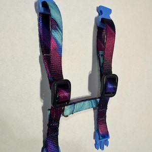 Puede incluir: Un arnés para mascotas con un patrón abstracto vibrante en tonos morado, azul y turquesa. El arnés presenta hebillas de plástico azul y clips de plástico negro. El diseño incluye una correa para el pecho y correas ajustables.