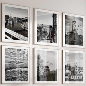 May include: A sectional sofa with beige and brown cushions, a set of three round dark metal coffee tables, and a light-coloured armchair. Six black and white framed photographs of cityscapes and landscapes are arranged on the wall above the sofa.