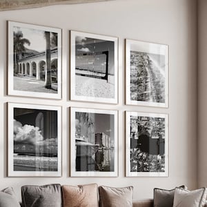 May include: A living room with a sectional sofa, a coffee table, and an armchair. The sofa is covered in a light brown fabric and has several pillows. The coffee table is made of wood and has a round top. The armchair is upholstered in a light gray fabric and has a gold metal frame. There are several black and white framed photographs on the wall behind the sofa.