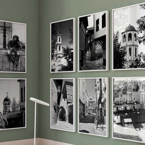 May include: A home office with a white desk and a white chair. The desk has three drawers and a laptop on top. There are eight black and white framed photos on the wall behind the desk. The photos are of buildings and streets in a city. There is a vase with dried flowers on the desk.