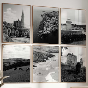 May include: A gallery wall set of six black and white framed prints featuring coastal landscapes, a historic church, a lighthouse, and a stone castle. The prints are arranged in two rows of three, displayed on a white wall above two beige armchairs.