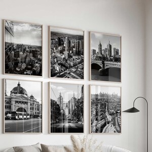 May include: A set of six black and white framed wall art prints featuring various cityscapes and architectural landmarks, arranged in a grid pattern on a white wall above a curved light gray sofa and a round white coffee table.