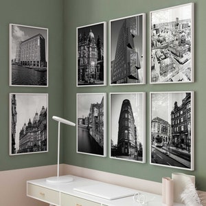 以下が含まれることがあります： 白い机に3つの引き出し、白い椅子、ランプ、ドライフラワーの入った花瓶が置かれたホームオフィス。壁は薄い緑色に塗られており、街並みの白黒写真が8枚額装されている。