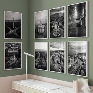 May include: A home office with a white desk and a white chair. The desk has three drawers and a laptop on top. There are six black and white framed photos of a city skyline on the wall behind the desk. There are two vases with dried flowers on the desk.