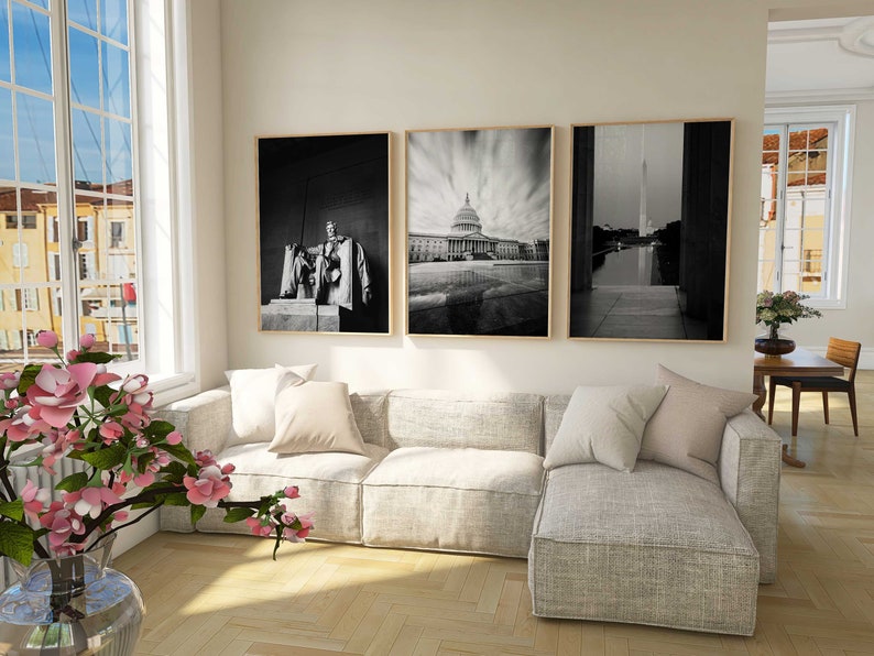 May include: Three black and white framed photographs of Washington D.C. landmarks. The photos are hung on a white wall above a light gray sectional sofa. The sofa has a light gray throw pillow and a white throw pillow.