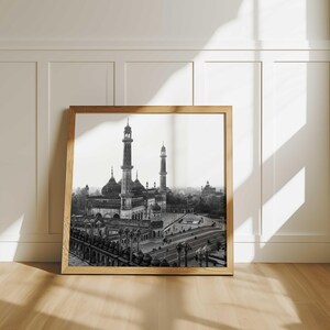 May include: Black and white framed print of a large mosque with tall minarets and domes. The image is set against a white wall with a wooden frame, and the floor is made of wood.
