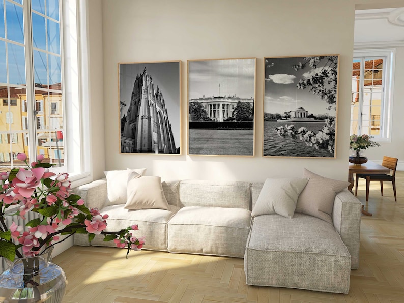 May include: A living room interior with a sectional sofa, three black and white framed photographs on the wall, and a vase of pink flowers on the floor.