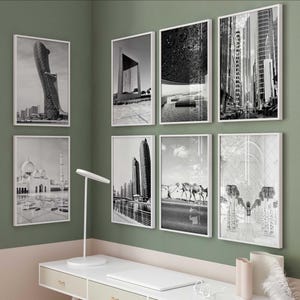 May include: A collection of eight framed black and white photographs of architectural landmarks and cityscapes. The images include a mosque, skyscrapers, and desert scenes with camels. The frames are white, and the wall is painted a muted green.