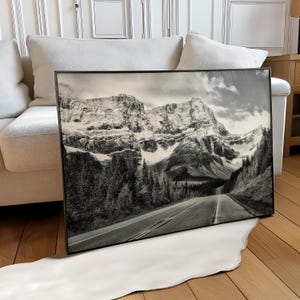 Peut inclure: Photographie encadrée en noir et blanc d'un paysage de montagne. Une route traverse une vallée vers des montagnes enneigées. L'image présente un ciel nuageux et des arbres à feuilles persistantes.