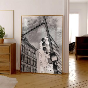 May include: Framed monochrome print featuring city architecture and a traffic light. The image shows a low-angle perspective of buildings and a traffic light against a cloudy sky. The buildings display detailed architectural elements.