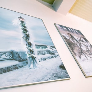 May include: Two framed photographs hanging on a white wall. The left photo shows a lamppost covered in snow and ice with a garland of greenery. The right photo shows a close-up of a deer's antlers in the snow.