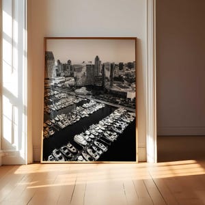 May include: Black and white framed print of a marina filled with boats, with a city skyline in the background. The print is displayed on a light-colored wooden floor, leaning against a white wall.