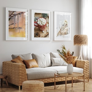 May include: Three framed winter-themed prints on a white wall above a wicker sofa with brown throw pillows. The prints feature a snowy owl, a cup of hot chocolate with marshmallows, and icicles hanging from a branch.