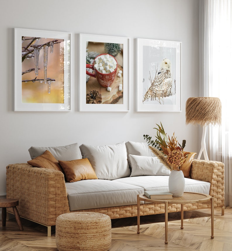 May include: Three framed winter-themed prints on a white wall above a wicker sofa with brown throw pillows. The prints feature a snowy owl, a cup of hot chocolate with marshmallows, and icicles hanging from a branch.