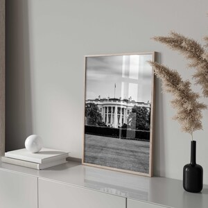 May include: A black and white photograph of the White House in Washington, D.C. The photo is framed in a light wood frame and is hanging on a white wall.