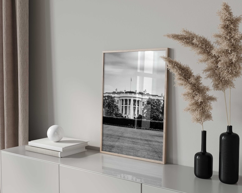 May include: A black and white photograph of the White House in Washington, D.C. The photo is framed in a light wood frame and is hanging on a white wall.