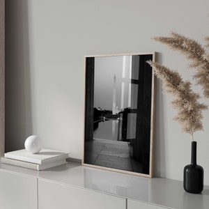 May include: A black and white photograph of a building with a tall spire, framed in a light wood frame. The photo is displayed on a white shelf with a white orb and two books.