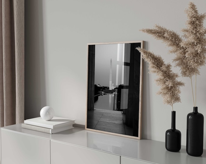 May include: A black and white photograph of a building with a tall spire, framed in a light wood frame. The photo is displayed on a white shelf with a white orb and two books.