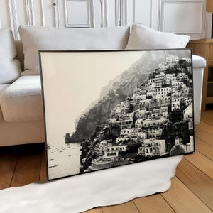 Op de afbeelding: Zwart-wit ingelijste prent van een Italiaanse kustplaats op een heuvel. De afbeelding toont gebouwen, een kerk en boten. De prent wordt gepresenteerd op een wit tapijt en leunt tegen een witte bank.