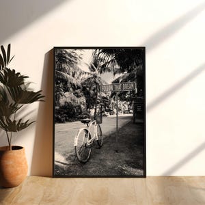 Puede incluir: Una impresión de arte mural enmarcada en blanco y negro que muestra una bicicleta aparcada en una carretera junto a una señal metálica que dice FOLLOW THAT DREAM. La escena presenta palmeras al fondo, creando un ambiente tropical.