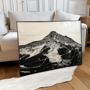 May include: Black and white framed print of a mountain landscape. The image features a tall mountain with snow-covered slopes and dark green trees. The print is displayed in a black frame.