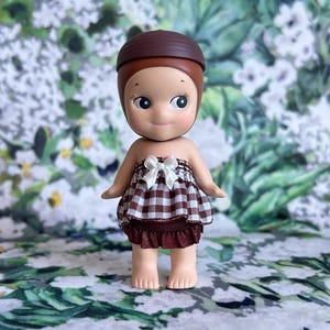 Puede incluir: Una pequeña figura con un sombrero marrón en forma de bellota y un vestido a cuadros con un lazo blanco. La figura tiene un tono de piel claro, mejillas rosadas y una expresión dulce. El fondo presenta un patrón floral.