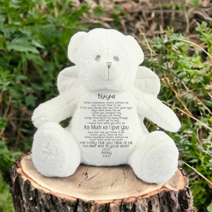 May include: A white teddy bear with angel wings, placed on a tree stump. The bear has a smiling face and a poem with the name "Blayne". The background is blurred green leaves.
