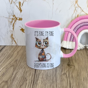 May include: A white ceramic mug with a pink rim and handle. The mug features a cartoon cat with a patchwork design and the text "It's fine, I'm fine, everything is fine."