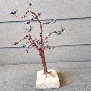 Op de afbeelding: Een handgemaakte draadboomsculptuur met een koperkleurige stam en takken. De takken zijn versierd met kleine, ronde, blauwe kralen, die op bessen lijken. De boom is gemonteerd op een rechthoekige, gebroken witte stenen basis.