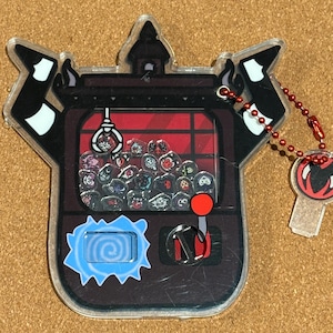 May include: A clear acrylic keychain shaped like a claw machine. The machine has a red interior with small character designs inside. The keychain has a red ball chain and a charm with a red and white design.
