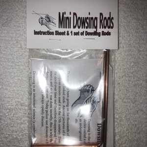 May include: A package containing a set of mini dowsing rods and an instruction sheet. The rods are made of copper and are bent at a 90-degree angle. The instruction sheet has the words "Mini Dowsing Rods" printed on it.