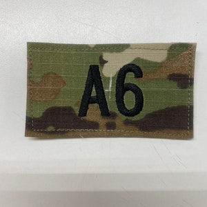 May include: A rectangular patch with a camouflage pattern in shades of green, brown, and tan. The patch features the embroidered text "A6" in black. The patch is likely for military or tactical use.