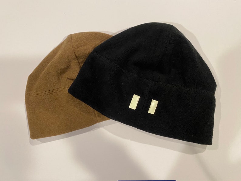 Puede incluir: Dos gorros de lana polar, uno marr&oacute;n y otro negro. El gorro negro tiene dos parches rectangulares blancos en la parte posterior.