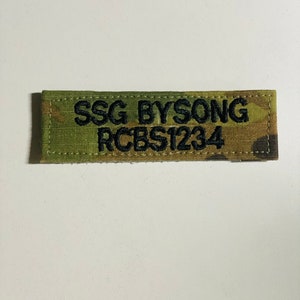 May include: A rectangular patch with a camouflage pattern. The patch has black embroidered text that reads "SSG BYSONG" above "RCBS1234". The edges of the patch are stitched with a matching thread.