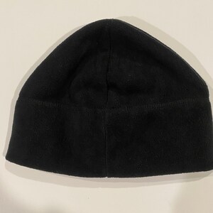 Puede incluir: Un gorro de punto negro de forro polar. El gorro est&aacute; doblado por la mitad y la costura es visible.