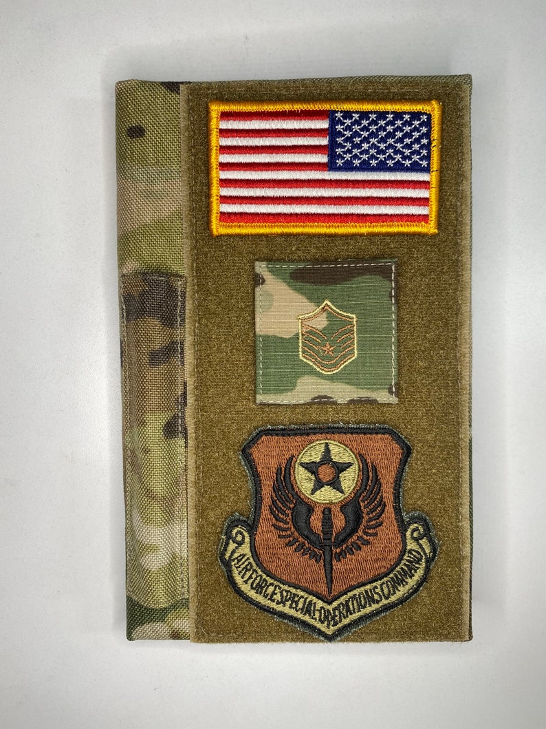 May include: A brown leather notebook cover with a camouflage pattern on the left side and a green fabric with patches on the right side. The patches include the American flag, a military rank patch, and a patch with the text "Air Force Special Operations Command".