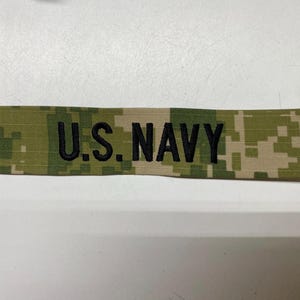 May include: A camouflage fabric patch with the text "U.S. NAVY" embroidered in black.