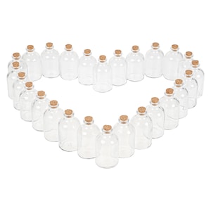 Puede incluir: Una forma de corazón hecha de 25 botellas de vidrio transparente con tapas de corcho. Las botellas están dispuestas en forma de corazón sobre un fondo blanco.