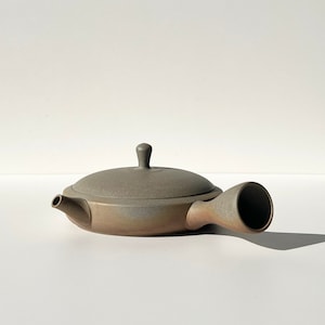 May include: A gray ceramic teapot with a rounded body and a long, curved spout. The teapot has a lid with a small knob on top.