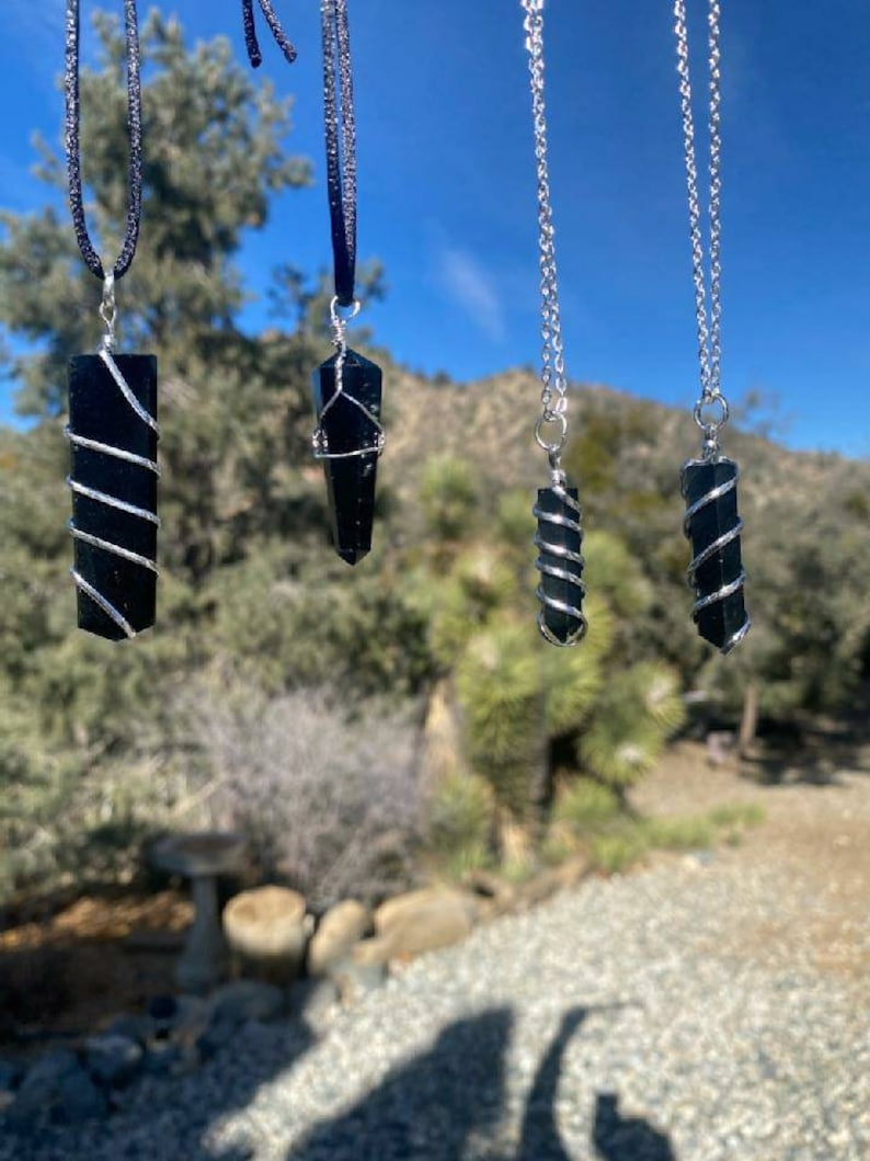 May include: Four black obsidian crystal pendants with silver wire wrapped around them, hanging from black and silver chains against a blue sky background.