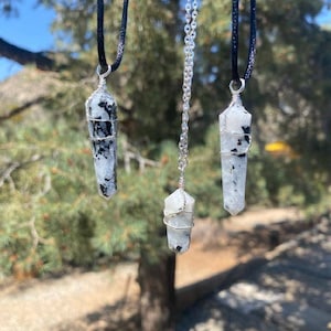 May include: Three silver wire wrapped crystal pendants on different chains. The crystals are white with black flecks and are shaped like long, thin points. The chains are black, silver, and black.