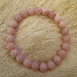May include: A light pink beaded bracelet. The bracelet is made of round, polished rose quartz beads. The beads are strung together to form a circular shape. The bracelet is displayed on a light-colored, textured surface.