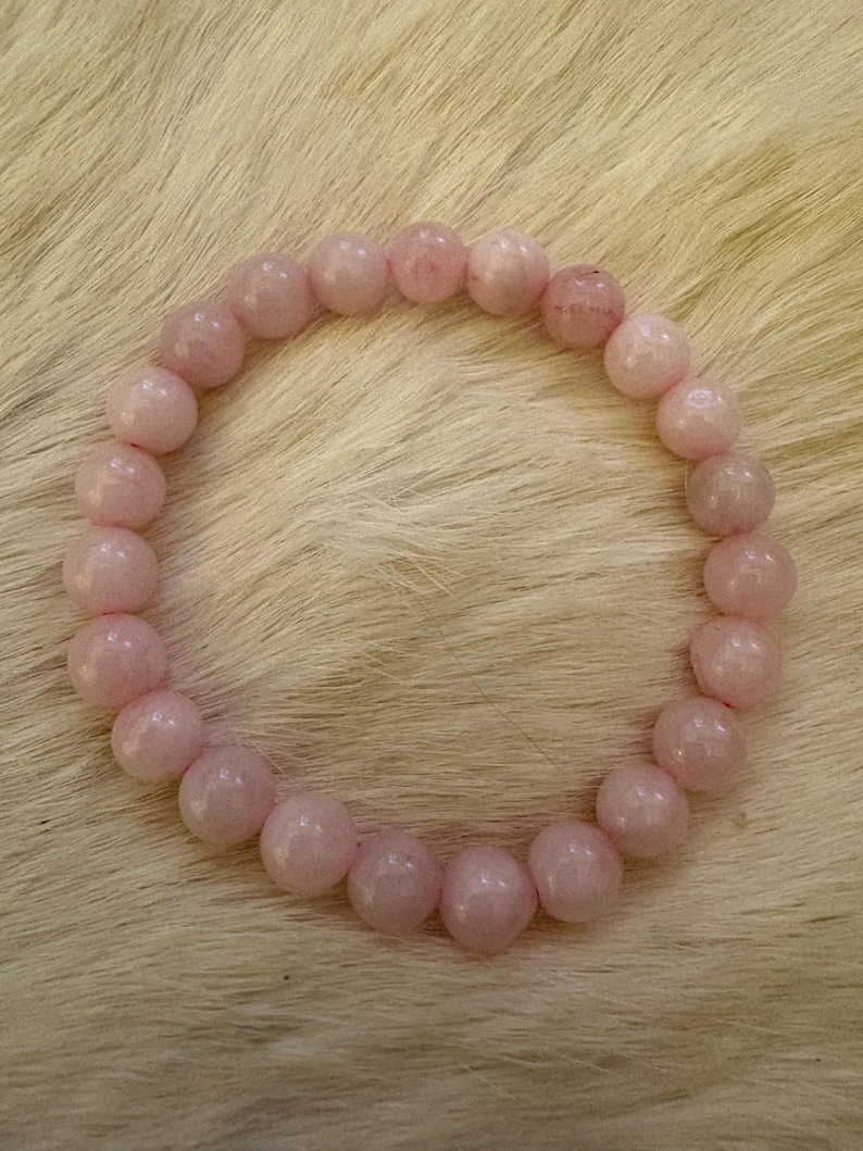May include: A light pink beaded bracelet. The bracelet is made of round, polished rose quartz beads. The beads are strung together to form a circular shape. The bracelet is displayed on a light-colored, textured surface.