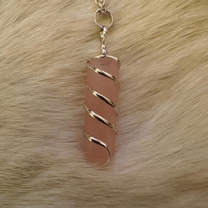 May include: A pink crystal pendant wrapped in silver-colored wire, suspended from a silver chain. The crystal is a rectangular shape. The background is a light-colored, textured surface.