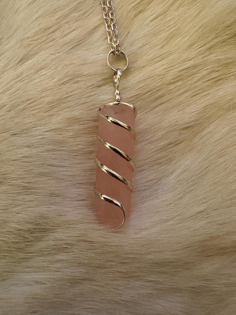 May include: A pink crystal pendant wrapped in silver-colored wire, suspended from a silver chain. The crystal is a rectangular shape. The background is a light-colored, textured surface.